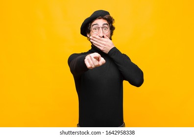 Young French Artist Man Laughing At You, Pointing To Camera And Making Fun Of Or Mocking You Against Orange Wall