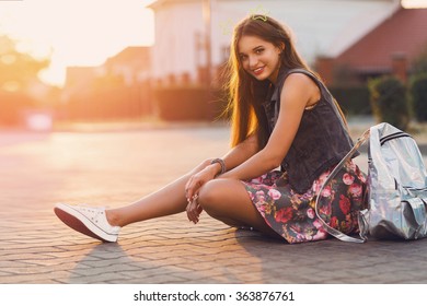 Young Fresh Cheerful Hipster  Girl  Sitting On The Road In Countryside Wearing Stylish Summer Clothes, Bright Print Skirt , Neon  Bag Pack. Warm Evening Colors. Sunset. Lifestyle Image.