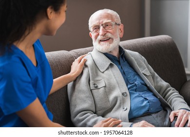 Young friendly african female caregiver talking chatting to happy senior man in hallway of nursing home. Picture of smiling nurse assisting senior elderly man