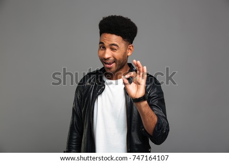 Young friendly african guy winks one eye while showing OK gesture, isolated on gray background