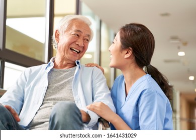 young friendly asian female caregiver talking chatting to happy senior man in hallway of nursing home