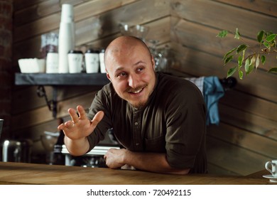 Young friendly bartender with mustache and beard standing at the bar counter in loft-styled cafe and meeting new customers with a smile. Former factory building, natural daylight. 