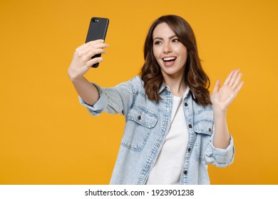 Young friendly fun woman 20s wearing stylish denim shirt white t-shirt doing selfie shot on mobile cell phone talking by video call waving hand greeting isolated on yellow background studio portrait.