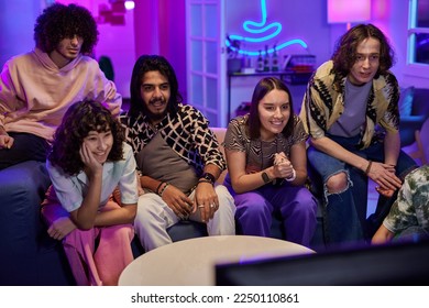 Young friendly intercultural people in casualwear watching tv set while sitting on comfortable couch in living room at home party - Powered by Shutterstock