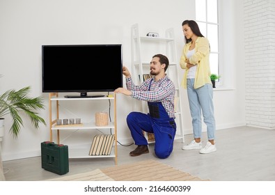 Young friendly service man installs and connects new TV in female client's house. Young woman watches as male repairman in work uniform connects cables to TV screen. Service and maintenance concept. - Shutterstock ID 2164600399