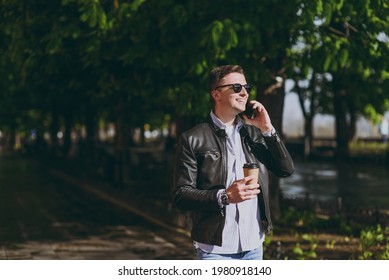 Young Friendly Stylish Man In Black Leather Jacket Eyeglasses Walk In Green Park Hold Takeaway Craft Paper Cup Drink Coffee To Go Talk By Mobile Cell Phone Concept Of Urban Lifestyle Spring Season.