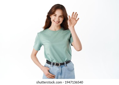 Young friendly woman waving hand and saying hello, greeting guests, making hi gesture and smiling broadly, standing against white background
