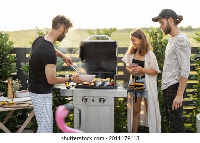 Young Friends Cooking Vegetables And Fish On A Modern Gas Grill At Backyard On A Sunset. Eating And Spending Summer Time Outdoors