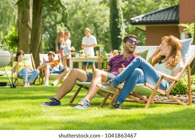 Young Friends Having Fun At Backyard Barbecue Party, Drinking Beer, Talking And Playing Guitar On Hot Summer Day, Focus On The Couple Laughing Sitting On Deck Chairs