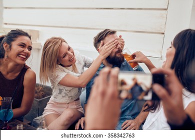 Young friends having fun at party being photographed with mobile phone. Young people sitting together enjoying party with one woman closing eyes of a man with another giving drink. - Powered by Shutterstock