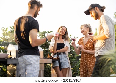 Young Friends Having Fun At Picnic Outdoors, Talk And Drink Wine At The Backyard On A Sunset. Spending Summer Time In A Small Group On The Open Air