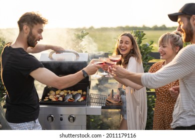 Young Friends Having Fun At Picnic Outdoors, Grilling Food And Drinking Wine At The Backyard On A Sunset. Spending Summer Time In A Small Group On The Open Air