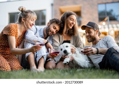 Young Friends Having Fun Playing With A Dog And Drinking Wine, Sitting On The Green Lawn At Backyard Of The Country House In The Evening