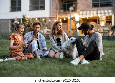 Young Friends Having Fun Playing With A Dog And Drinking Wine, Sitting On The Green Lawn At Backyard Of The Country House In The Evening