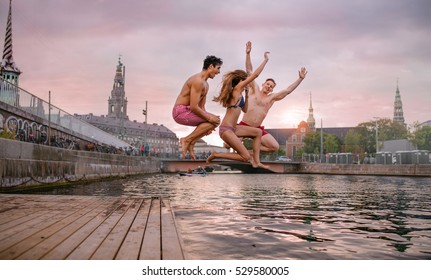 Young Friends Jumping From Jetty Into Lake. Group Of People In Mid Air At The Lake.