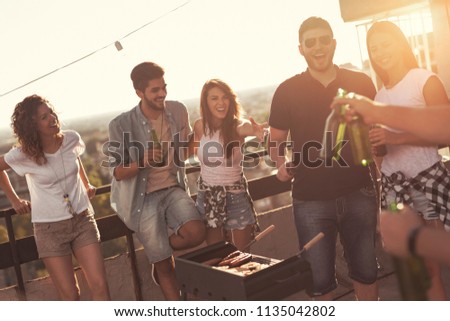 Young friends making barbecue, drinking beer and enjoying hot summer days having fun on a rooftop party. Focus on the guy next to the barbecue