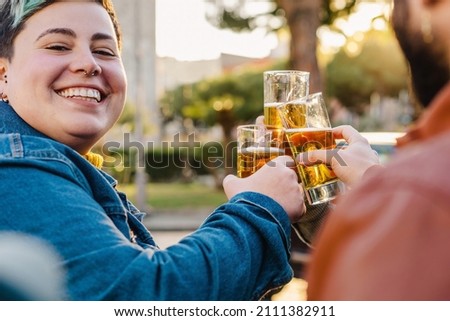 Young friends refresh themselves drinking ice-cold craft beer - sitting at the kiosk bar table a group of best friends celebrate by raising their glasses and making them clink - focus on glasses