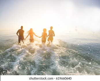 Young Friends Running Into The Sea At Sunset - Freedom And Vacation Concept - People Having Fun Inside Ocean Water On Summer Vacation With Back Sun Flare - Warm Filter With Soft Vignette