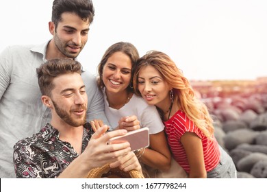 Young friends sitting outdoors and looking at smartphone. Young man showing something interesting to friends outdoors at the seaside - Powered by Shutterstock