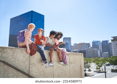 Young friends sitting and using smart phones, hanging out on ledge in sunny city