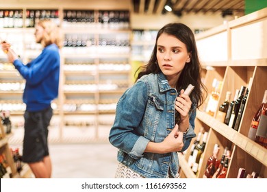 Young frightened woman in denim jacket trying steal bottle of wine in modern supermarket