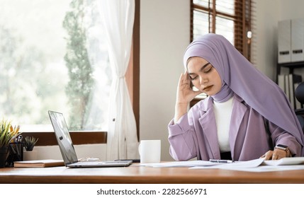 Young frustrated arabic Muslim young woman in hijab working at office desk in front of laptop suffering from chronic daily headaches - Shutterstock ID 2282566971