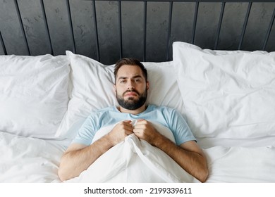 Young Frustrated Despressed Sad Unshaven Unhappy Man Wear Blue T-shirt Lying In Bed Look Camera Rest Relax Spend Time In Bedroom Lounge Home In Own Room House Wake Up. Bad Mood Night Bedtime Concept