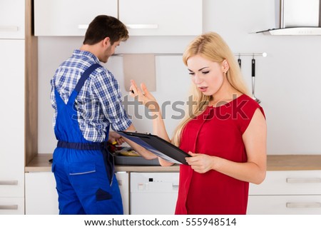 Young Frustrated Woman Looking At Expensive Bill On Clipboard With Plumber Standing In Kitchen