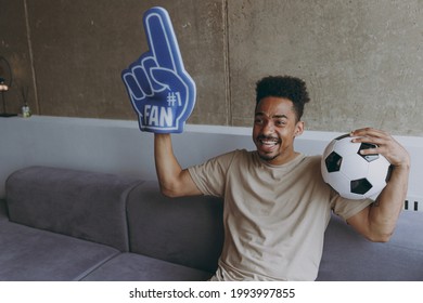 Young Fun African American Man In Beige T-shirt Cheer Up Support Favorite Team With Soccer Ball Football Fan Foam Glove Finger Up Shout Sit On Grey Sofa Indoors Apartment People Sport Leisure Concept