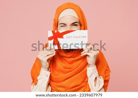 Young fun arabian asian muslim woman wear orange abaya hijab hold gift certificate coupon voucher card for store blinkeye isolated on plain pink background. Uae middle eastern islam religious concept