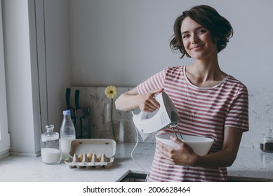 Young fun cheerful brunette housewife woman 20s in casual clothes striped t-shirt using mixer whips yolks beats eggs cooking food in light kitchen at home alone. Healthy diet bakery lifestyle concept