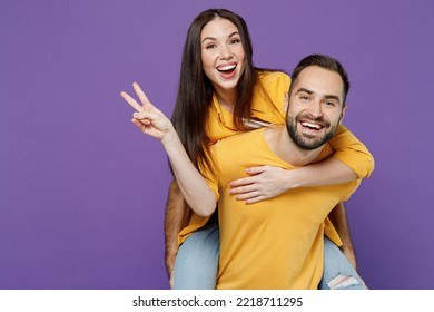 Young fun cheerful couple two friends family man woman together in yellow casual clothes giving piggyback ride to joyful, sit on back show v-sign isolated on plain violet background studio portrait.