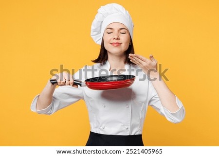 Young fun chef cook baker woman wear white shirt black apron uniform toque chefs hat hold frying pan raised hand to face, feeling food smell isolated on plain yellow background. Cooking food concept