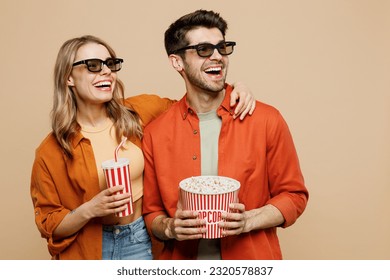 Young fun couple two friends family man woman in 3d glasses wear casual clothes watch movie film hold bucket of popcorn cup of soda pop in cinema look aside isolated on plain beige background studio