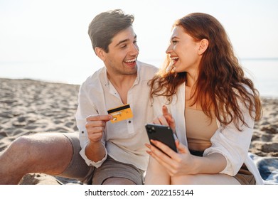 Young Fun Couple Two Man Woman In White Clothes Sit On Sand Use Mobile Phone Credit Card Shopping Online Order Delivery Book Tour Rest Together At Sunrise Over Sea Beach Outdoor Seaside In Summer Day