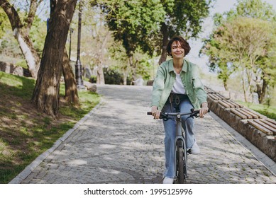 Young fun dreamful happy woman 20s wearing casual green jacket jeans riding bicycle bike on sidewalk in city spring park outdoors, look aside. People active urban healthy lifestyle cycling concept. - Shutterstock ID 1995125996