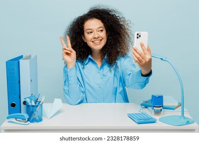 Young fun employee business woman wearing casual shirt sit work at white office desk doing selfie shot on mobile cell phone show v-sign isolated on plain pastel light blue background studio portrait