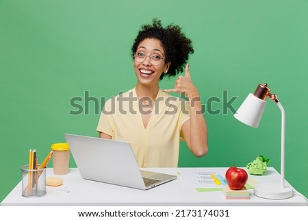 Young fun employee business woman of African American ethnicity wear shirt sit work at white office desk with pc laptop doing phone gesture like says call me back isolated on plain green background.