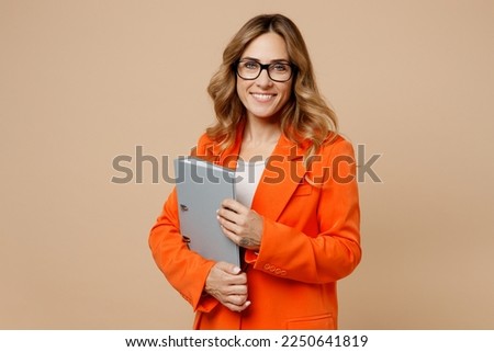 Young fun employee business woman corporate lawyer 30s wear classic formal orange suit glasses work in office hold grey folder for papers document bookkeeping isolated on plain beige background studio