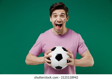 Young fun european man fan supporter wears basic pink t-shirt cheer up support football sport team hold in hand soccer ball watch tv live stream scream isolated on dark green color background studio