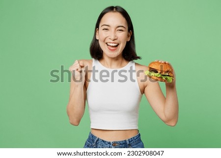 Young fun excited woman wear white clothes hold eat burger do winner gesture clench fist isolated on plain pastel light green background. Proper nutrition healthy fast food unhealthy choice concept