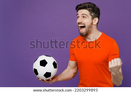 Young fun exultant happy fan man he 20s wearing orange t-shirt cheer up support football sport team hold in hand soccer ball watch tv live stream do winner gesture isolated on plain purple background
