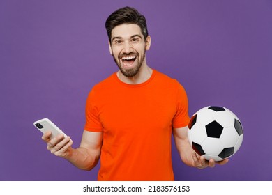 Young fun fan happy cheerful man he wears orange t-shirt cheer up support football sport team hold in hand soccer ball watch tv live stream use mobile cell phone isolated on plain purple background - Shutterstock ID 2183576153