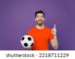 Young fun fan man he wear orange t-shirt cheer up support football sport team hold in hand soccer ball watch tv live stream point index finger overhead on workspace isolated on plain purple background