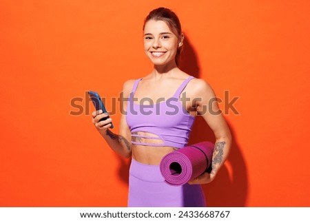 Young fun fitness trainer instructor sporty woman sportsman wear purple top clothes in home gym hold yoga mat use mobile cell phone isolated on plain orange background. Workout sport fit abs concept