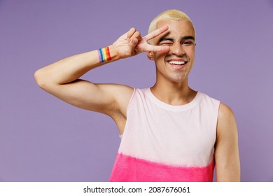 Young fun friendly latin gay man 20s with make up wearing bright pink top cover eye with victory sign isolated on plain pastel purple background studio portrait. People lifestyle fashion lgbtq concept