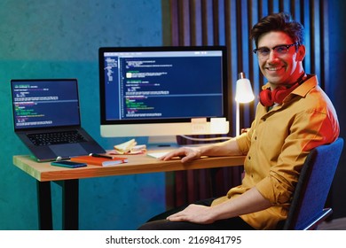 Young Fun Happy Back-end Developer Software Engineer IT Specialist Programmer Man In Shirt Headhones Work At Home Writing Code On Laptop Pc Destop Computer Analyzing Data. Program Development Concept