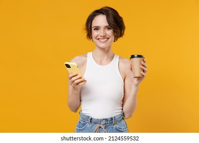 Young fun happy caucasian smiling woman 20s with bob haircut wearing white tank top shirt hold takeaway delivery craft paper brown cup coffee to go, mobile cell phone isolated on yellow background. - Powered by Shutterstock