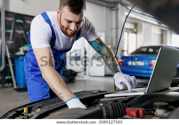 Young fun happy professional technician car\
mechanic man in blue overalls t-shirt use laptop pc computer make\
diagnostics check fix problem with raised hood work in vehicle\
repair shop workshop\
indoor