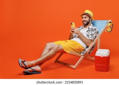 Young fun happy tourist man in beach shirt hat lie on deckchair near fridge hold use mobile cell phone isolated on plain orange background studio portrait. Summer vacation sea rest sun tan concept. - Shutterstock ID 2154835891